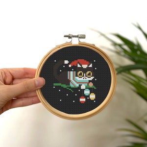 May include: A black cat wearing a red Santa hat and a blue and white scarf is holding a red and white ornament. The cat is sitting on a green branch with white snow falling around it. The image is a cross-stitch pattern on black fabric in a wooden embroidery hoop.