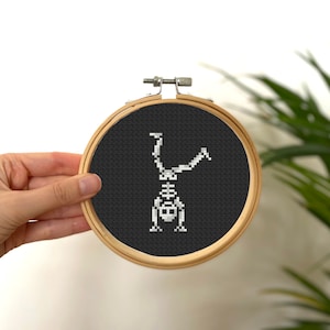 May include: A white skeleton cross-stitched on black fabric in a wooden embroidery hoop. The skeleton is doing a handstand.