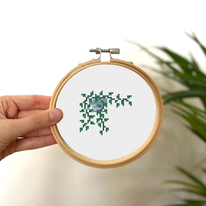 May include: A cross-stitch embroidery hoop with a green vine design. The vine is stitched in a light green color and has a small blue flower.