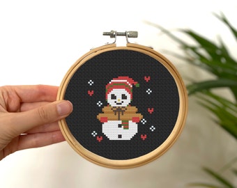 May include: A black and white cross-stitch embroidery of a snowman wearing a red and green hat and scarf. The snowman is surrounded by red hearts and white snowflakes.