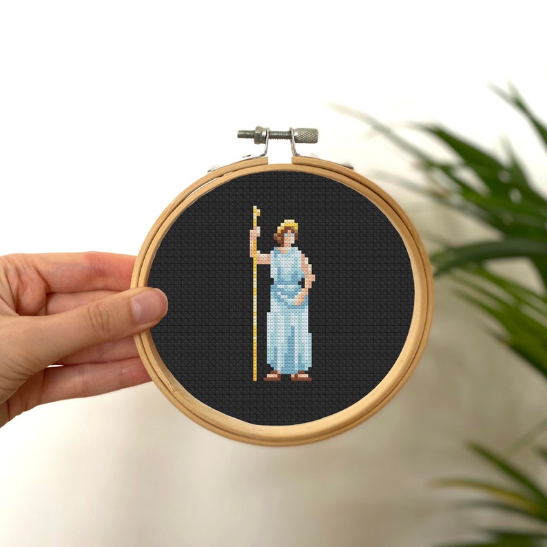 May include: A cross-stitch embroidery of a woman in a white robe holding a staff, framed in a wooden hoop. The background is black.