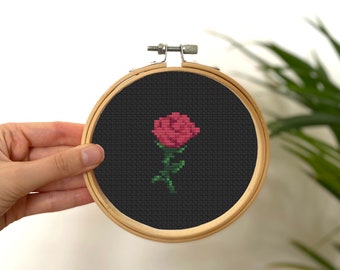 May include: A small, black embroidery hoop with a pink rose cross-stitched on a black background.