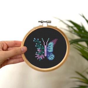 Puede incluir: Un aro de bordado negro con un diseño de punto de cruz de una mariposa púrpura y azul con follaje verde.