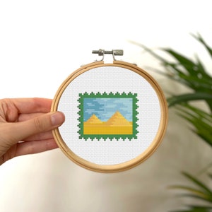 Puede incluir: Un aro de bordado de punto de cruz con un marco verde y una imagen de un paisaje desértico con dos pirámides amarillas y un cielo azul.