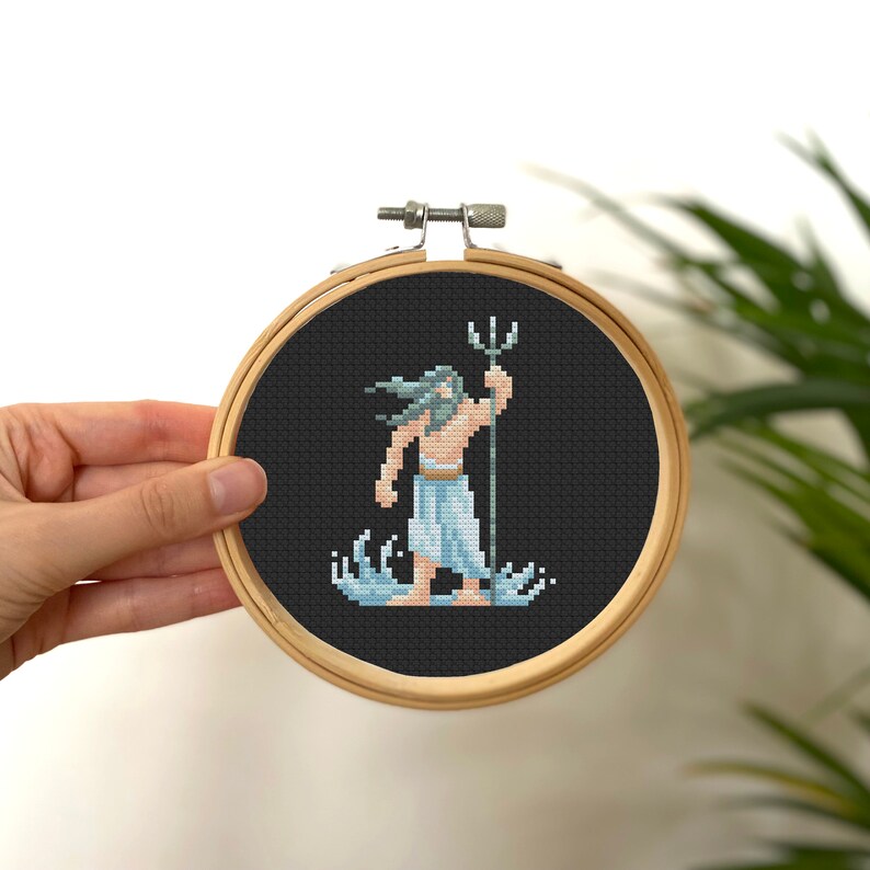 May include: A cross-stitch embroidery hoop with a black background and a colorful pixelated image of Poseidon, the Greek god of the sea, holding a trident. He is standing in the water with waves around him.