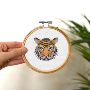 May include: A close-up of a cross-stitch embroidery of a tiger's face in a wooden embroidery hoop. The tiger has orange and black stripes, a pink nose, and white fur around its eyes.