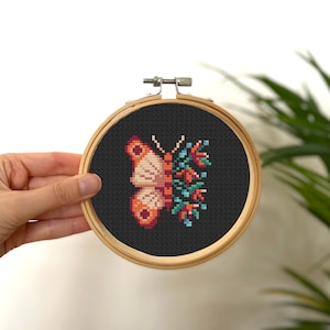 Könnte beinhalten: Ein Kreuzstich-Stickrahmen mit schwarzem Hintergrund, auf dem ein weißer und orangefarbener Schmetterling mit floralen Details abgebildet ist.