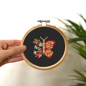 Könnte beinhalten: Eine Kreuzstich-Stickerei eines Schmetterlings mit floralen Details auf schwarzem Hintergrund. Der Schmetterling ist orange, gelb und grün. Die Stickerei ist in einem Holzrahmen eingefasst.