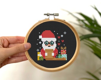 May include: A cross-stitch embroidery hoop with a black background and a pixelated image of Santa Claus wearing glasses and holding a gift. The image is surrounded by snowflakes and hearts.