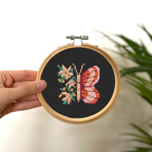May include: A cross-stitch embroidery of a butterfly with orange and pink wings, surrounded by white flowers with green leaves, on a black background. The embroidery is framed in a wooden hoop.