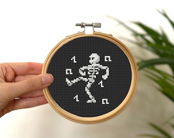 May include: A black and white cross-stitch embroidery hoop with a pixelated skeleton dancing on a black background.