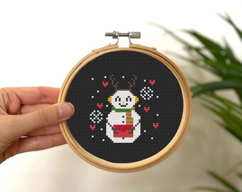 May include: A cross-stitch embroidery hoop featuring a snowman design. The snowman has antlers, a green scarf, and a red container. The background is black, and the hoop is held in a hand.