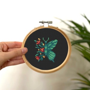 以下が含まれることがあります： 蝶と花の模様のクロスステッチ刺繍枠。蝶はティールとグリーンで、オレンジと赤の花があります。背景は黒で、枠は木製です。