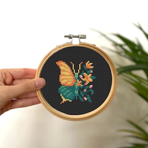 Könnte beinhalten: Eine Kreuzstich-Stickerei eines Schmetterlings mit orangefarbenen und gelben Flügeln, grünem und blauem Körper und rosa Blüten auf schwarzem Hintergrund. Die Stickerei ist in einem Holzrahmen eingefasst.