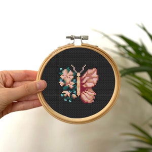 Könnte beinhalten: Kreuzstich-Stickerei eines Schmetterlings und Blumen in einem Holzreifen. Der Schmetterling hat rosa und pfirsichfarbene Flügel, mit einem dunkelgrauen Hintergrund. Die Blumen sind rosa und blau.