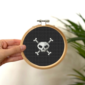 May include: A white skull and crossbones design embroidered on black fabric in a wooden embroidery hoop.