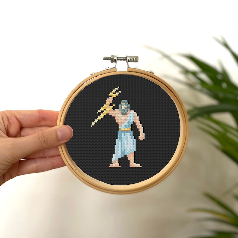 May include: A cross-stitch embroidery of Zeus, the Greek god of the sky and thunder, holding a lightning bolt. The embroidery is framed in a round wooden hoop and is set against a black background.