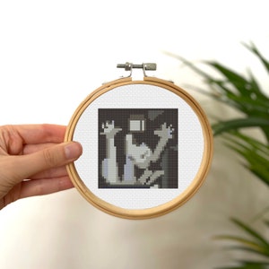 May include: A cross-stitch embroidery hoop with a black and white pixelated image of a person with their arms raised in the air.