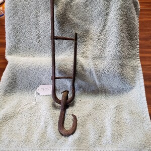 May include: A rusty, antique metal hook with a long handle and a smaller, ladder-like structure. The hook is curved and appears to be made of iron. The item is resting on a light green towel. A tag with the number 7037 is attached.