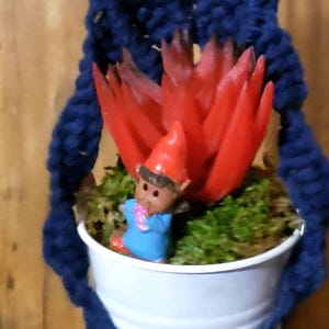 May include: A handmade macrame plant hanger in navy blue, holding a white pot with a red succulent and a small gnome figurine. The tag above reads "9044".