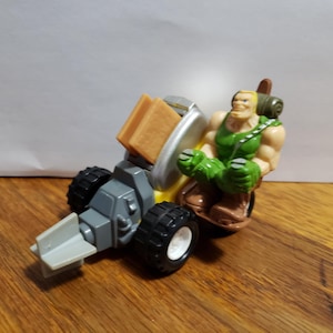 May include: A toy vehicle with a muscular action figure. The vehicle is gray, yellow, and black with white wheels. The action figure is wearing a green tank top and brown boots. The figure has a backpack and is smiling.