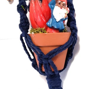 May include: A navy blue macrame plant hanger with a small terracotta pot. Inside the pot is a red and green decorative arrangement and a gnome figurine. The hanger has a wooden bead and a tassel.
