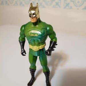May include: A green Batman action figure with a gold-colored cowl and belt. The figure has black gloves and boots. The Batman logo is on the chest. The figure is standing.