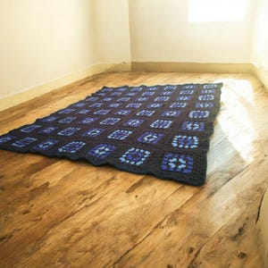 May include: A handmade, dark blue crochet blanket with a repeating square pattern in shades of blue. The blanket is displayed on a wooden floor, showcasing its texture and design. The edges have a scalloped finish.