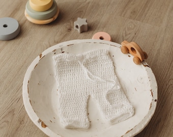 May include: A pair of white crocheted pants with a drawstring waist, placed inside a distressed white wooden bowl. Nearby are wooden toys including a stacking ring set, a star-shaped block, and a small wooden car.