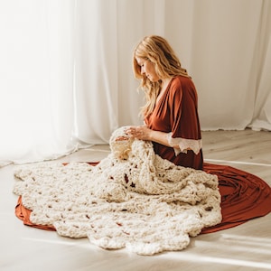 May include: A chunky, cream-coloured crocheted blanket featuring a repeating floral or shell-like pattern. The blanket is crafted from thick, soft yarn and displays a textured, open-work design with scalloped edges throughout the entire piece.