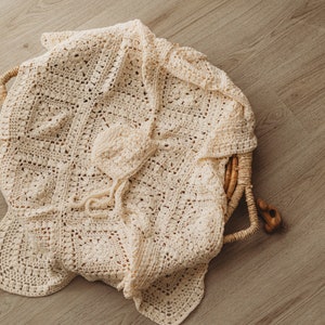 May include: A cream-colored crocheted baby blanket with a matching crocheted hat. The blanket is folded and placed in a woven basket.