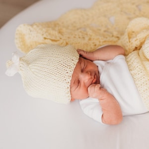 Può includere: Un neonato con un berretto di lana giallo e un body bianco sta dormendo su una coperta di uncinetto gialla.