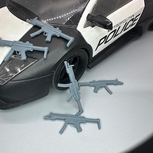 May include: Five grey plastic toy machine guns, some lying on a white surface and some on a black and white toy police car with the words "Metropolitan Police" on the side.