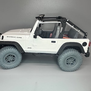 Puede incluir: Coche de juguete Jeep Rubicon blanco con ruedas negras y una jaula antivuelco negra. El coche tiene un logotipo "Jeep" negro en el lateral y "Rubicon" escrito en el capó.
