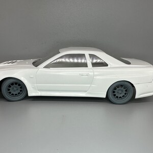 May include: White plastic model car of a Nissan Skyline GT-R R34, a popular Japanese sports car. The model is in a side profile view, showing the car's sleek lines and distinctive features.