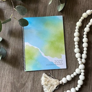 May include: A 2027 monthly planner with a spiral binding and a watercolor-style cover featuring blended shades of blue and lime green. The text 2027 MONTHLY PLANNER is printed in the bottom right corner in a clean, dark sans-serif font.