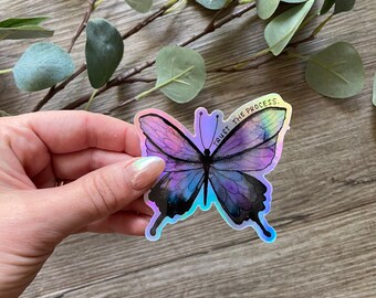 May include: A holographic sticker featuring a purple and blue butterfly with the text "TRUST THE PROCESS."