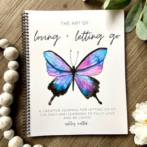 Peut inclure: Un journal à spirales avec un papillon aquarelle sur la couverture. Le titre du journal est "The Art of Loving + Letting Go". Le sous-titre est "A creative journal for letting go of the past and learning to fully love and be loved." Le nom de l'auteur est "ashley mattek".