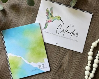 May include: A 2027 spiral-bound monthly planner with a blue and green watercolor cover and a 2027 wall calendar featuring a watercolor hummingbird illustration and the text 2027 Calendar 12 Months of Whimsical Well Wishes for the Soul.