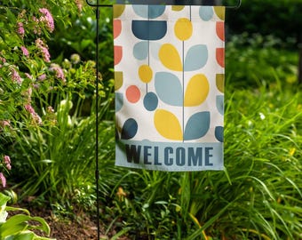 May include: A garden flag with a colorful, abstract design of yellow, blue, and orange shapes on a cream background. The word "WELCOME" is printed in blue at the bottom of the flag. The flag is displayed outdoors in a garden.