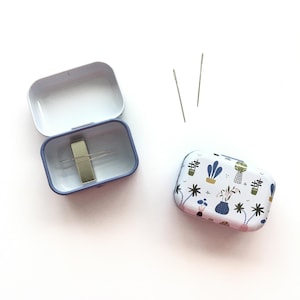 May include: A small, rectangular sewing needle tin with a white background. The tin is open, revealing needles and a magnetic holder. A second, closed tin with a botanical pattern is also present, along with two loose needles.