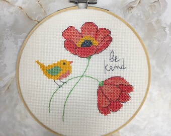 May include: A cross-stitch embroidery hoop with a white background, featuring a yellow bird perched on a green stem with two red poppy flowers. The text "be kind" is stitched in black thread.