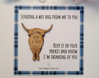 Può includere: Una spilla in legno a forma di mucca delle Highlands con le parole "A Wee Hug" su una carta con un bordo a quadri blu. La carta recita "Sending a wee hug from me to you. Keep it in your pocket and know I'm thinking of you."