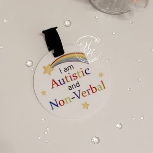 May include: A white, circular tag with a black strap. The tag displays a rainbow, stars, and the text "I am Autistic and Non-Verbal" in multiple colours. The Hidden Heart logo is also visible.