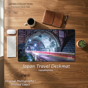 Può includere: Un tappetino da scrivania rettangolare con una stampa fotografica di un tunnel con scie luminose, etichettato Japan Collection Premium Desk Mat e Japan Travel Deskmat Inakalifephoto, posizionato su una superficie in legno con tastiera e mouse.