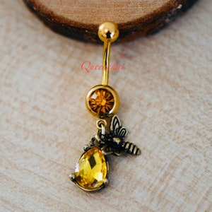 May include: Gold belly button ring with a dangling charm featuring a bee and a teardrop-shaped yellow crystal. The charm is attached to a smaller round yellow crystal.