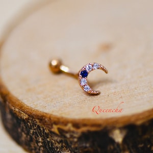 May include: A gold crescent moon body jewelry with a blue gemstone and clear rhinestones.