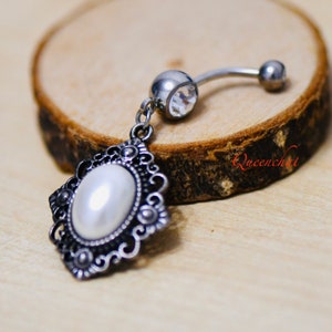 May include: A silver body jewellery piece with a clear crystal and a dangling ornate charm with a white pearl. The charm has a filigree design.