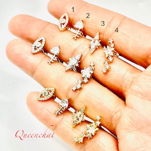 May include: Eight pairs of silver and gold earrings with clear crystal accents. The earrings feature various designs, including an evil eye, a star, a crescent moon, and a leaf. The earrings are numbered 1 through 8.