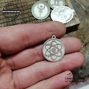 May include: A silver pendant with a detailed atom design. The pendant is 925 sterling silver and gold plated. The pendant is held in a hand.